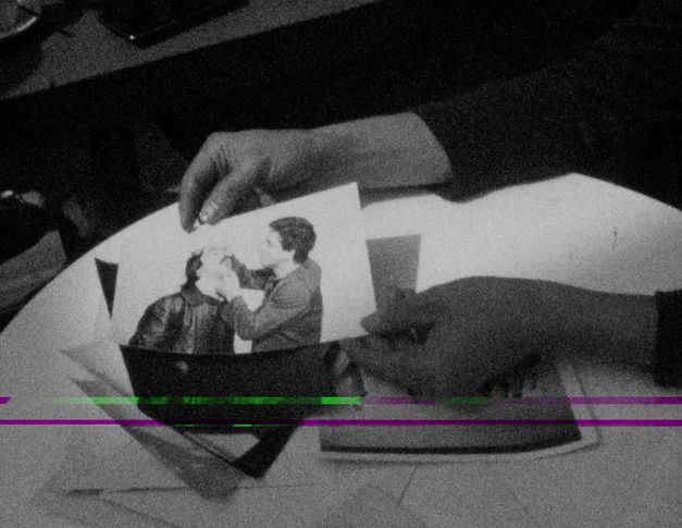 Filmstill aus „This Makes Me Want to Predict the Past“ von Cana Bilir-Meier. Nahaufnahme von zwei Händen, die ein Foto in der Hand halten. Auf dem Foto sieht man zwei Männer, von denen der eine den Kopf des anderen am Kinn und an der Stirn hält und nach hinten beugt. 