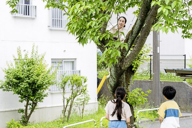 A woman in a white blouse climbs a tree. In front of the tree are two children watching her. In the background is a white panel building. 