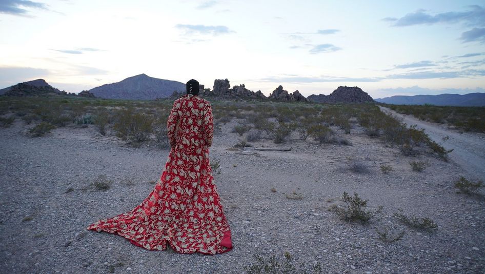 Filmstill aus CASSANDRO, THE EXOTICO: Man sieht von hinten eine Person, die in einer wüstenähnlichen Landschaft steht. Die Person trägt einen roten, überlangen, aufwendig verzierten Mantel.