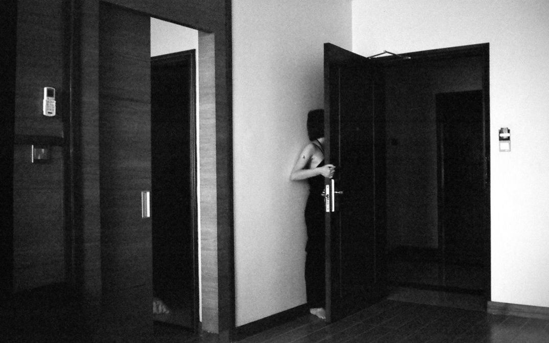 Still from the film "Jet Lag" by Zheng Lu Xinyuan. A black-and-white image of a girl hiding behind an open hotel room door, with the side of her body and her hair visible.