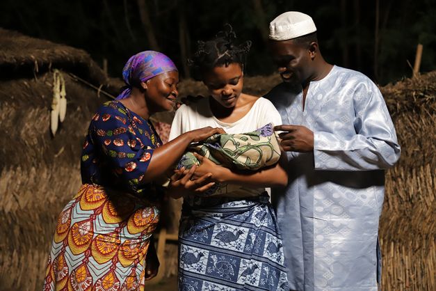 Filmstill aus dem Film „CERTAIN WINDS FROM THE SOUTH“ von Eric Gyamfi. Drei Personen lächeln, während sie ein in eine Decke gehülltes Baby halten, ihre Augen strahlen Wärme und Zärtlichkeit aus.