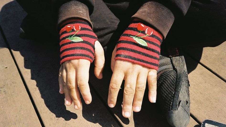 Film still from HAUBI: A person is sitting cross-legged on the floor. All you can see are their black shoes and their hands, which are wearing red, fingerless gloves.