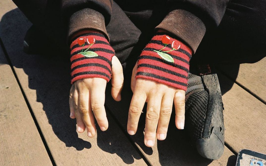 Filmstill aus HAUBI: Eine Person sitzt im Schneidersitz am Boden. Man sieht nur ihre schwarzen Schuhe und die Häne, die in roten, fingerlosen Handschuhen stecken.