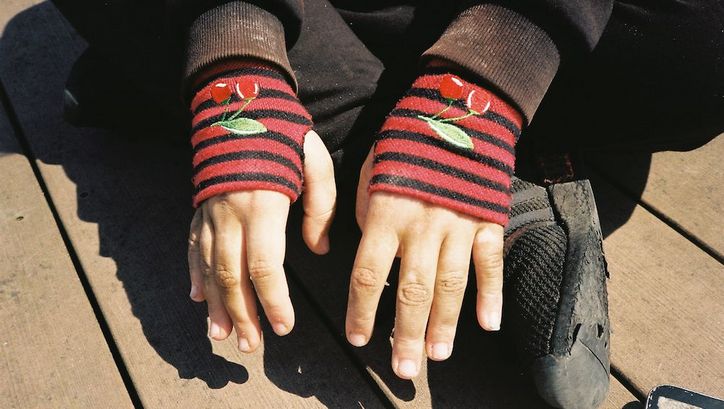 Filmstill aus HAUBI: Eine Person sitzt im Schneidersitz am Boden. Man sieht nur ihre schwarzen Schuhe und die Häne, die in roten, fingerlosen Handschuhen stecken.