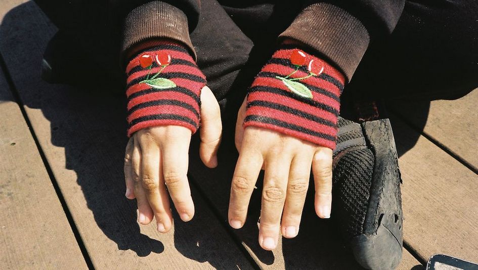 Filmstill aus HAUBI: Eine Person sitzt im Schneidersitz am Boden. Man sieht nur ihre schwarzen Schuhe und die Häne, die in roten, fingerlosen Handschuhen stecken.