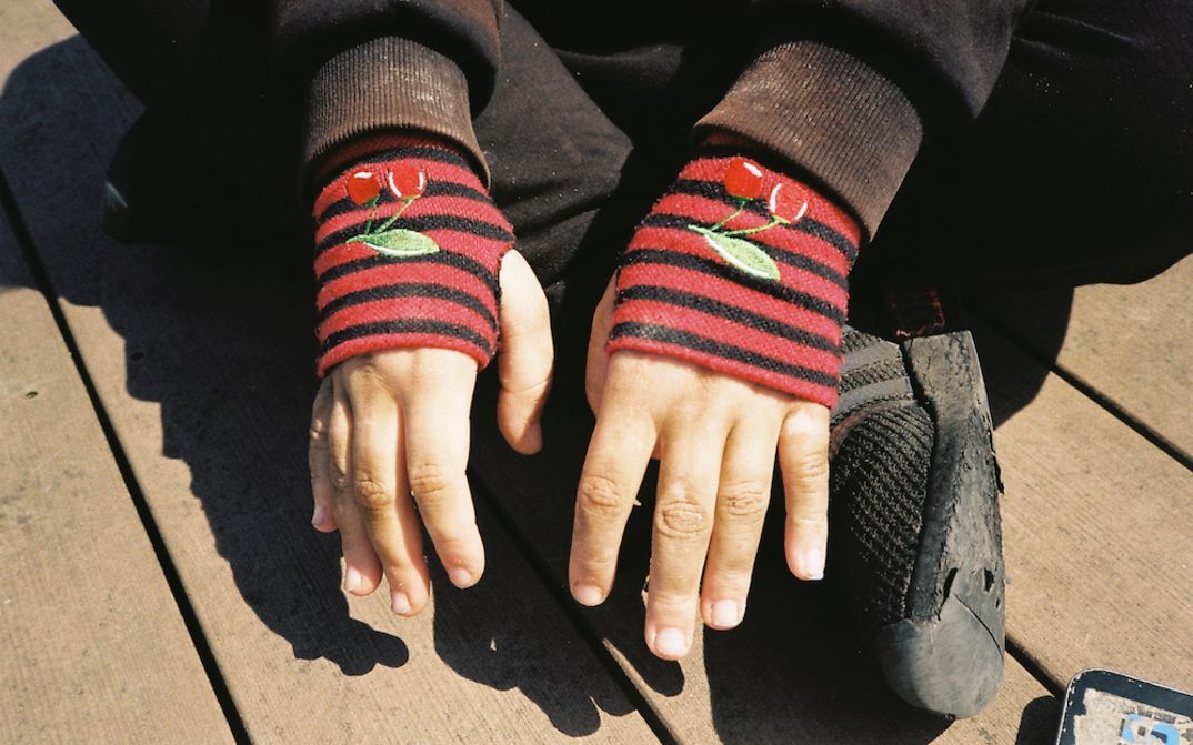 Filmstill aus HAUBI: Eine Person sitzt im Schneidersitz am Boden. Man sieht nur ihre schwarzen Schuhe und die Häne, die in roten, fingerlosen Handschuhen stecken.
