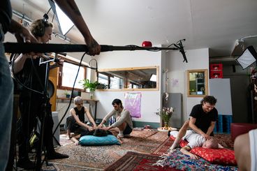 Eine Hinter-den-Kulissen-Aufnahme mit sieben Personen. Fünf von ihnen sitzen und liegen auf dem Boden, während die beiden anderen den Ton aufnehmen.