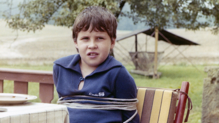 Filmstill aus VERSTECKTE FALLEN: Ein gefesselter Junge sitzt an einem Gartentisch.