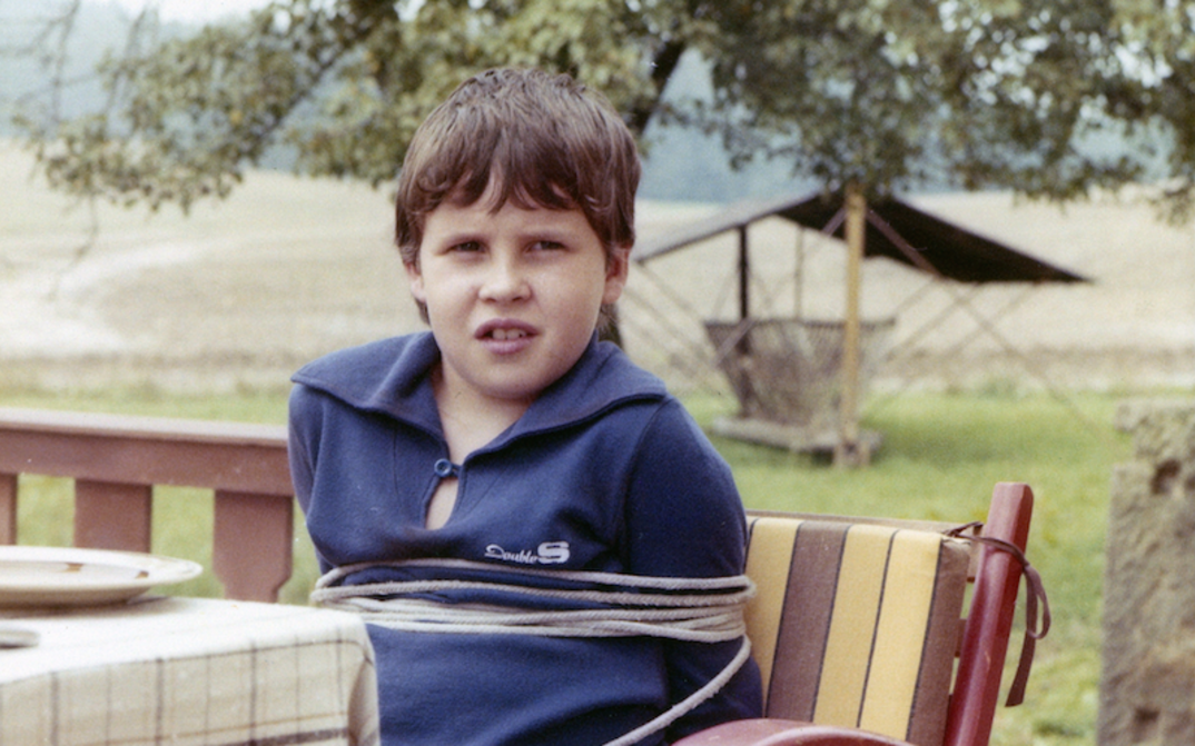 Film still from VERSTECKTE FALLEN: A tied-up boy sits at a garden table.