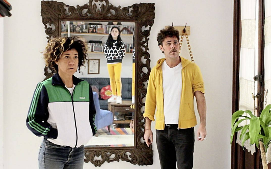 Still from the film "La edad media (The Middle Ages)" by Alejo Moguillansky and Luciana Acuña. A man and a woman stand next to each other in a living room, in the mirror behind them we see the reflection of their daughter standing on a coffee table. 