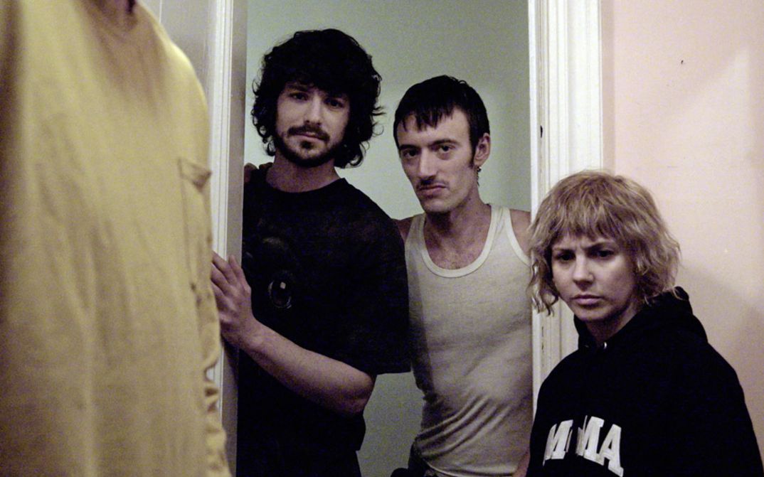 Film still from "The Wrong Movie" by Keren Cytter. It shows three people standing in the doorway and looking towards the camera. A fourth person is standing at the front left of the picture. Only the t-shirt of fourth person is visible.