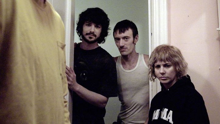Film still from "The Wrong Movie" by Keren Cytter. It shows three people standing in the doorway and looking towards the camera. A fourth person is standing at the front left of the picture. Only the t-shirt of fourth person is visible. 