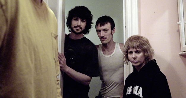 Film still from "The Wrong Movie" by Keren Cytter. It shows three people standing in the doorway and looking towards the camera. A fourth person is standing at the front left of the picture. Only the t-shirt of fourth person is visible. 
