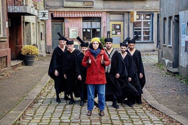 Film still from "Shahid" by Narges Kalhor. It shows a group of people between the houses on the street. In front is a woman in a red jacket. Behind her is a group of men dressed in black. 