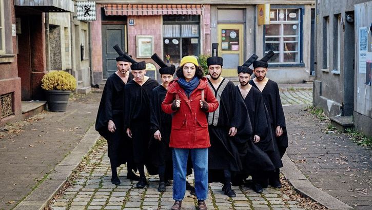 Film still from "Shahid" by Narges Kalhor. It shows a group of people between the houses on the street. In front is a woman in a red jacket. Behind her is a group of men dressed in black. 