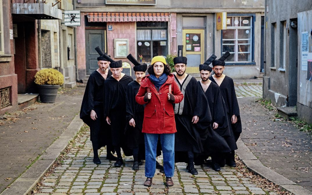 Filmstill aus "Shahid" von Narges Kalhor. Zu sehen ist eine Gruppe von Menschen zwischen den Häusern auf der Straße. Vorne steht eine Frau in einer roten Jacke. Hinter ihr steht eine Gruppe in Schwarz gekleideter Männer.