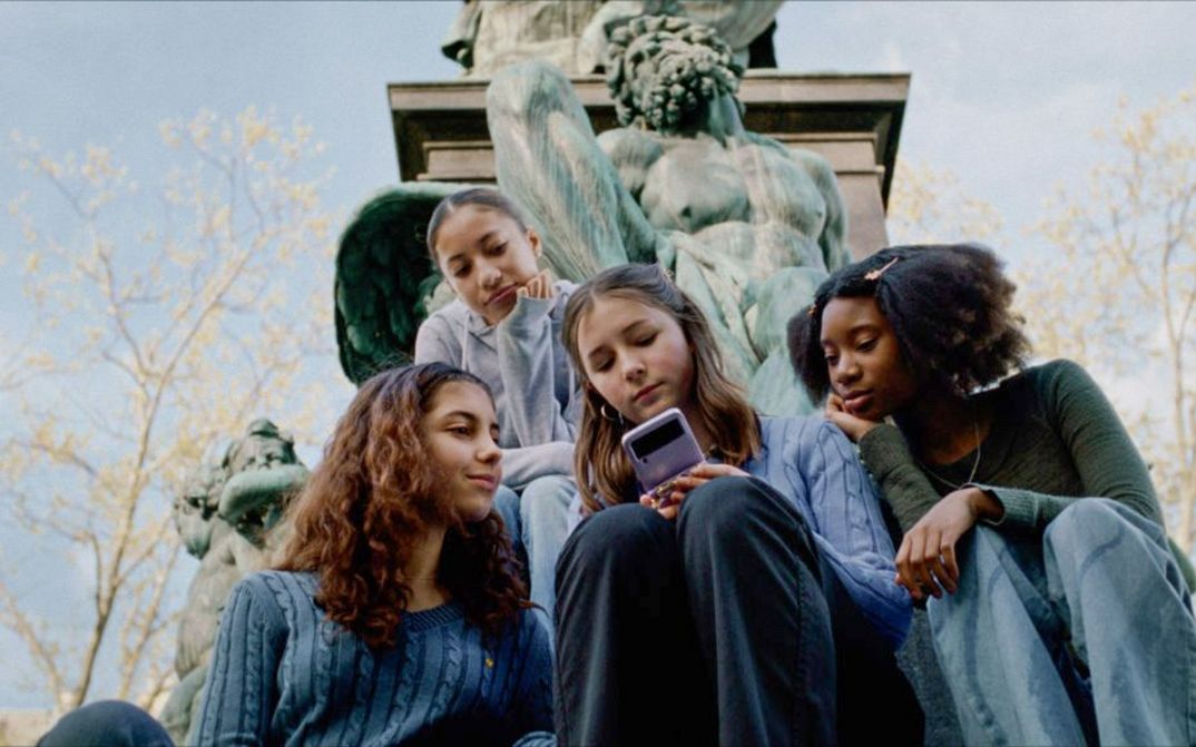 Filmstill aus WENN DU ANGST HAST NIMMST DU DEIN HERZ IN DEN MUND UND LÄCHELST von Marie Luise Lehner.  Vier junge Frauen sitzen vor einer Statue, alles schauen auf das Handy, das die mitlere von ihnen in der Hand hält.