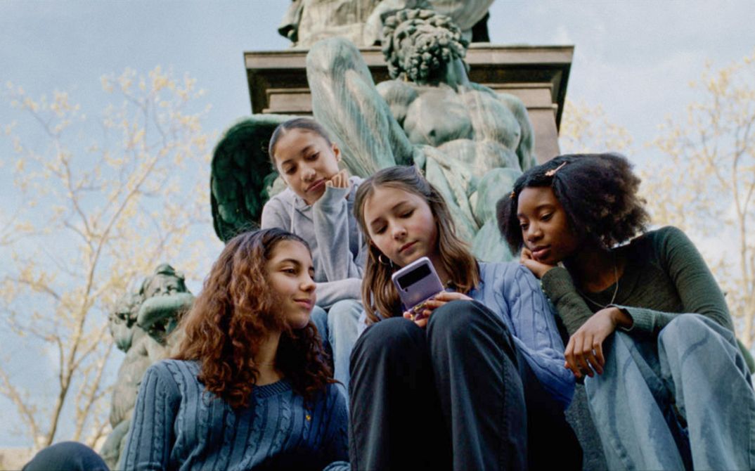 Filmstill aus WENN DU ANGST HAST NIMMST DU DEIN HERZ IN DEN MUND UND LÄCHELST von Marie Luise Lehner. Vier junge Frauen sitzen vor einer Statue, alles schauen auf das Handy, das die mitlere von ihnen in der Hand hält.