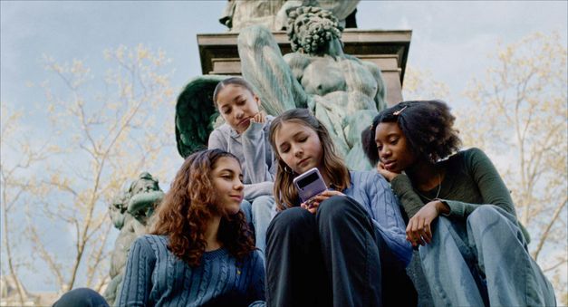  Film still from WENN DU ANGST HAST NIMMST DU DEIN DEIN HERZ IN DEN MUND UND LÄCHELST by Marie Luise Lehner. Four young women are sitting in front of a statue, all looking at the cell phone that the middle one is holding. 