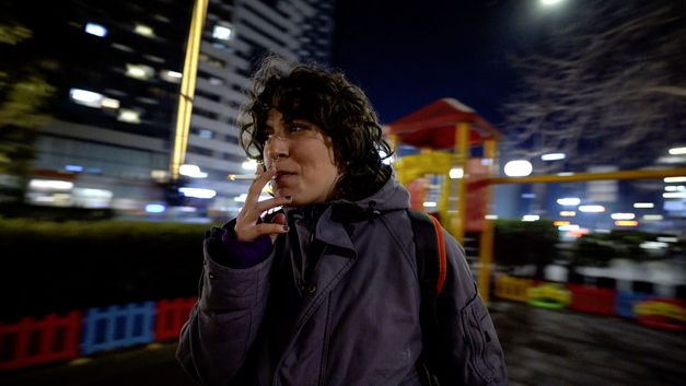 Still from INNER BLOOMING SPRINGS by Tiku Kobiashvili. The picture shows a woman smoking in front of a city backdrop at night.