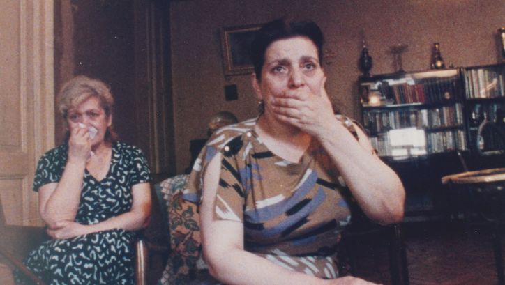 Film still from AM RANDE DER WELT: Two women are sitting in a living room, watching television. One is holding her hand over her mouth in shock, while the other is holding a handkerchief to her face.