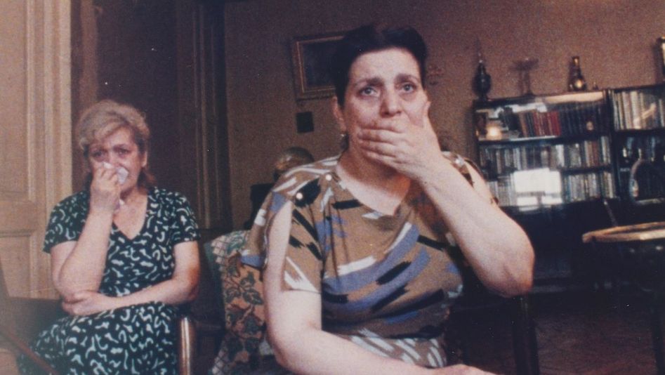 Filmstill aus AM RANDE DER WELT: Zwei Frauen sitzen in einem Wohnzimmer und blicken auf einen Fernseher. Eine hält sich erschrocken die Hand vor den Mund, die andere hält ein Taschentuch vor ihr Gesicht.