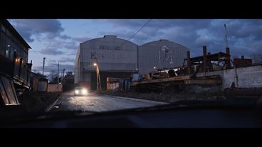 A white factory building with the words "Astilleros Mestrina" at the face of the building, at dawn, in front of it a car with headlights on drives towards the camera.
