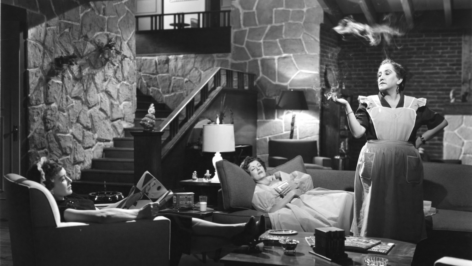 Film still from EL CASO DE LA MUJER ASESINADITA: Two women are sitting and lying on the couch in a living room. A maid stands in the middle of the room smoking.