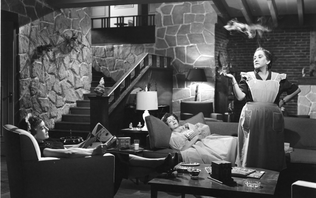 Filmstill aus EL CASO DE LA MUJER ASESINADITA: In einem Wohnzimmer sitzen und liegen zwei Frauen auf der Couch. Ein Dienstmädchen steht rauchend mitten im Raum.