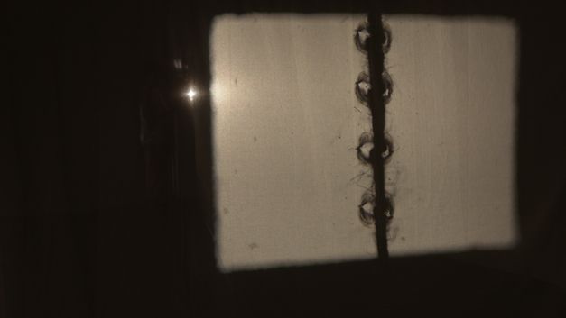 Film still from "The Song of the Shirt" by Kerstin Schroedinger. A screen lit from behind with a shadow of an indefinable object.