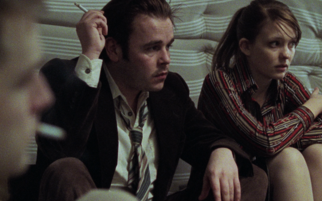 Filmstill aus BAADER: Zwei junge Menschen sitzen nebeneinander auf einer Matratze. Eine weitere Person ist angeschnitten und unscharf zu sehen.