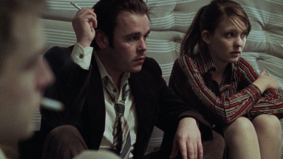 Filmstill aus BAADER: Zwei junge Menschen sitzen nebeneinander auf einer Matratze. Eine weitere Person ist angeschnitten und unscharf zu sehen.