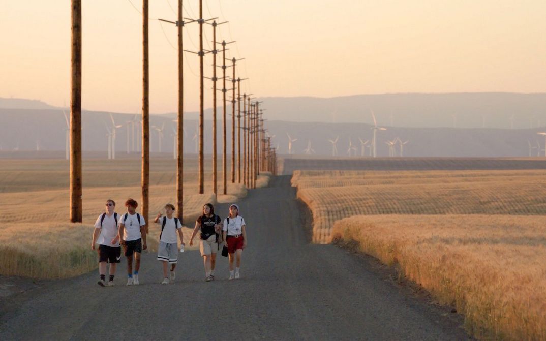 A group of teenagers walk along a slightly hilly and empty road between cornfields in the soft evening light.Film still from GASOLINE RAINBOW: