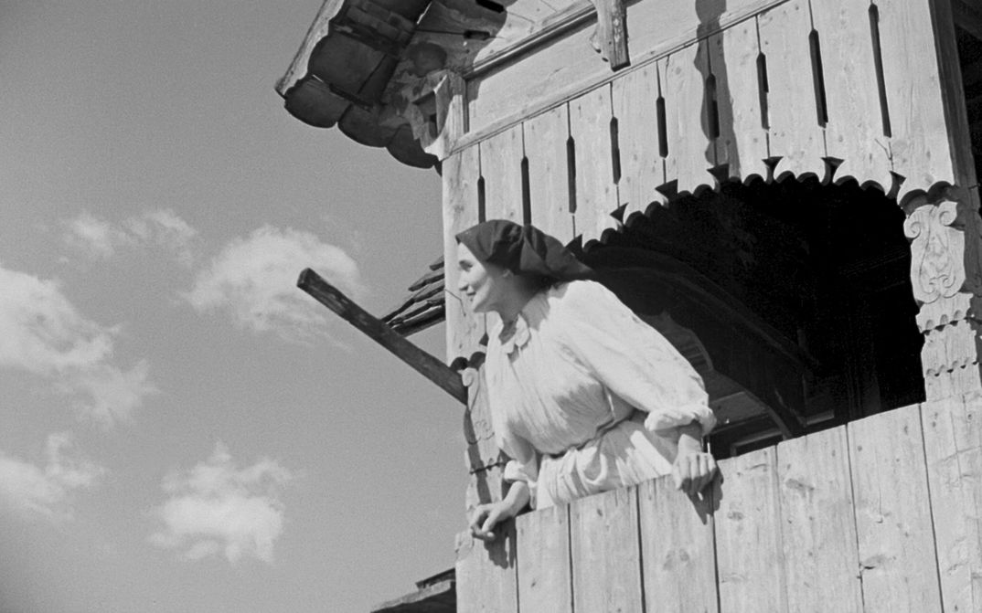 Film still from A Day in the Life of a Turopolje Peasant Community: A woman in a white dress and a black head scarf is looking through the window of a wooden building into the sky.