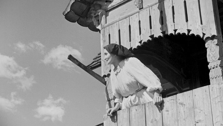 Film still from A Day in the Life of a Turopolje Peasant Community: A woman in a white dress and a black head scarf is looking through the window of a wooden building into the sky.