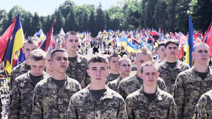 Soldiers stand in the sunlight. Ukrainian flags in the background. 