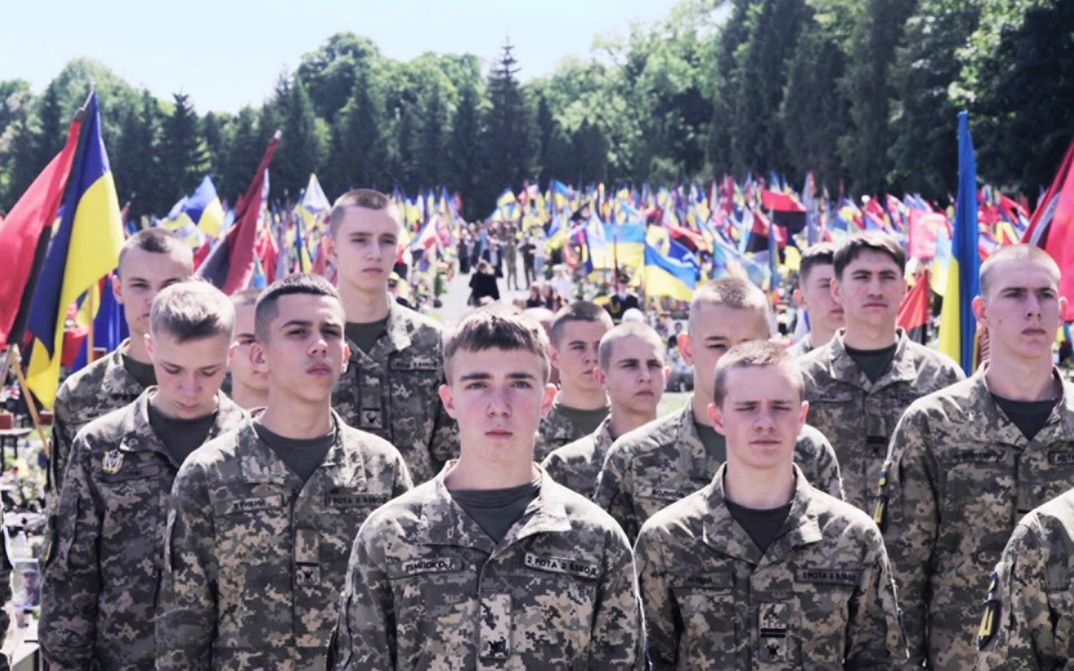 Soldiers stand in the sunlight. Ukrainian flags in the background.