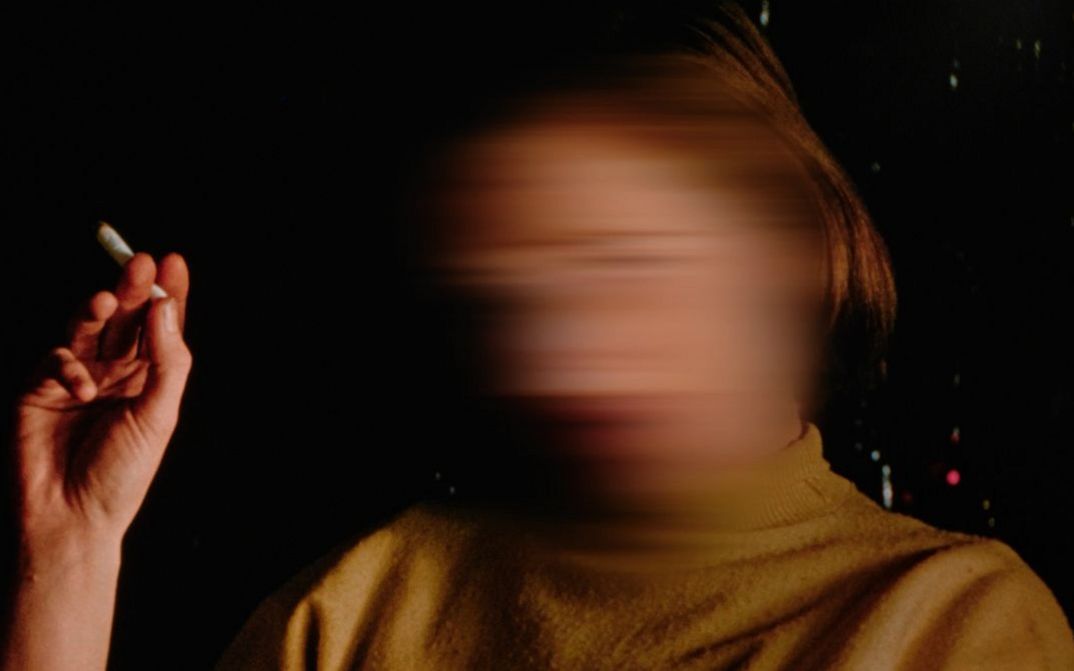 Filmstill aus dem Film "Home When You Return" von Carl Esaesser. Eine rauchende Frau mit verwischtem Gesicht vor dunklem Hintergrund.