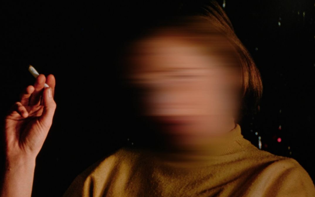 Filmstill aus dem Film "Home When You Return" von Carl Esaesser. Eine rauchende Frau mit verwischtem Gesicht vor dunklem Hintergrund.