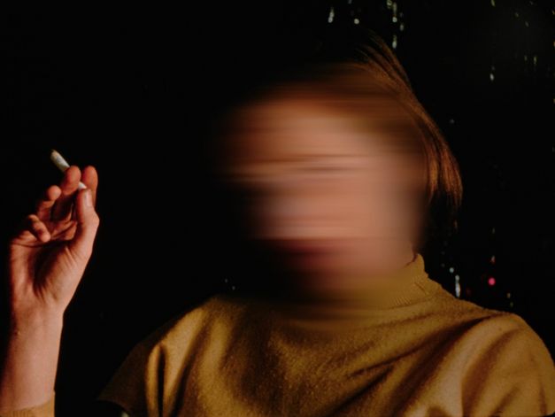 Filmstill from the film "Home When You Return" by Carl Esaesser. A smoking woman with a blurred face in front of a dark background.
