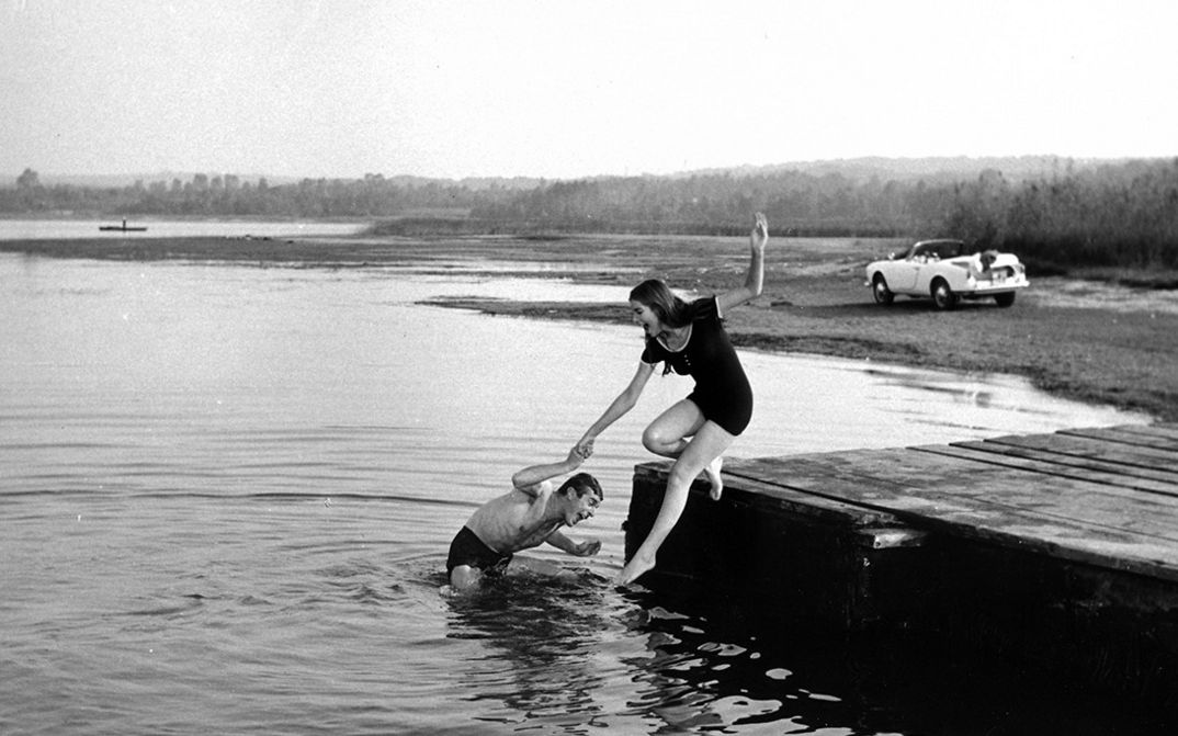 Film still from NATA DI MARZO: A man and a woman in bathing clothes at the edge of a body of water. He stands in the water and pulls her in. 