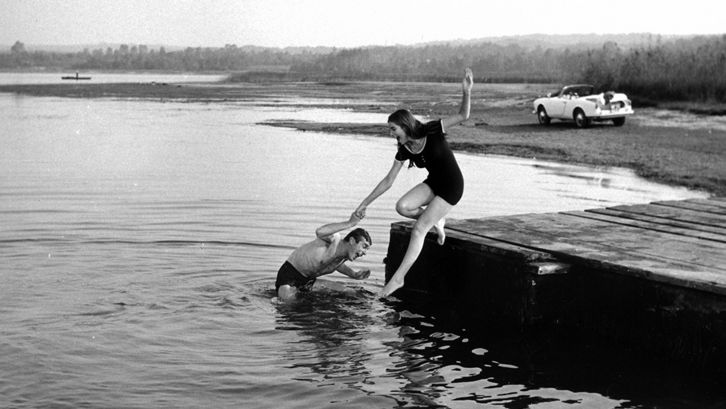 Film still from NATA DI MARZO: A man and a woman in bathing clothes at the edge of a body of water. He stands in the water and pulls her in. 