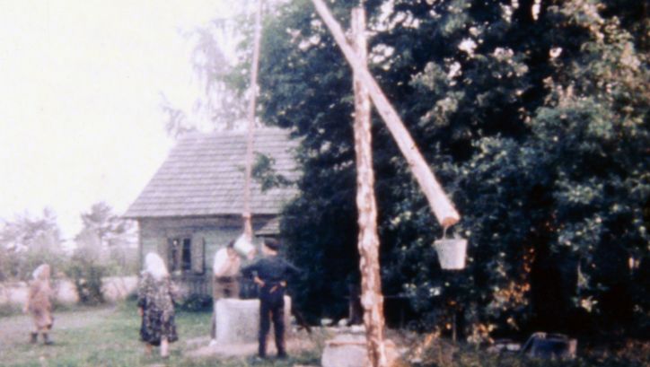 Film still from REMINISCENCES OF A JOURNEY TO LITHUANIA: Some people in a garden with a well, in the background a simple house.