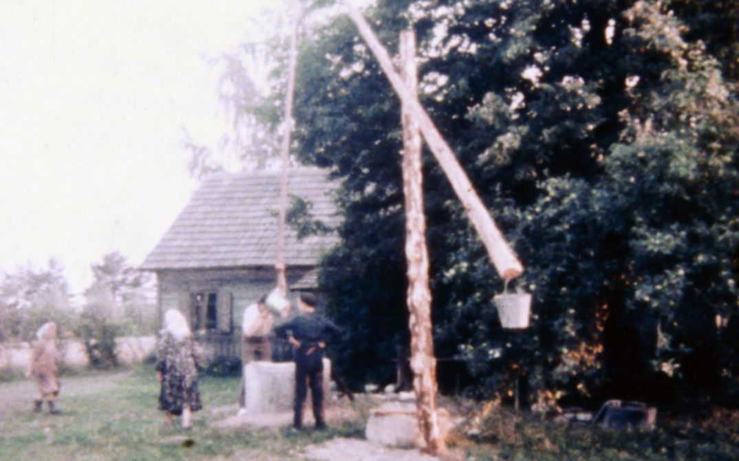 Film still from REMINISCENCES OF A JOURNEY TO LITHUANIA: Some people in a garden with a well, in the background a simple house.