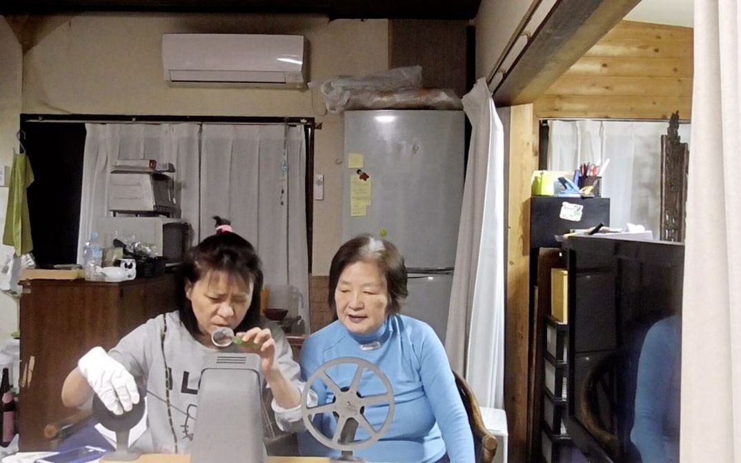 Filmstill aus "Voices of the Silenced" von Park Soo-nam und Park Maeui. Zu sehen sind zwei Frauen, die an einem Tisch in einem Raum mit einer Küche sitzen. Die Frau links legt einen Film in eine Spule ein. Die Frau auf der rechten Seite beobachtet sie dabei. 