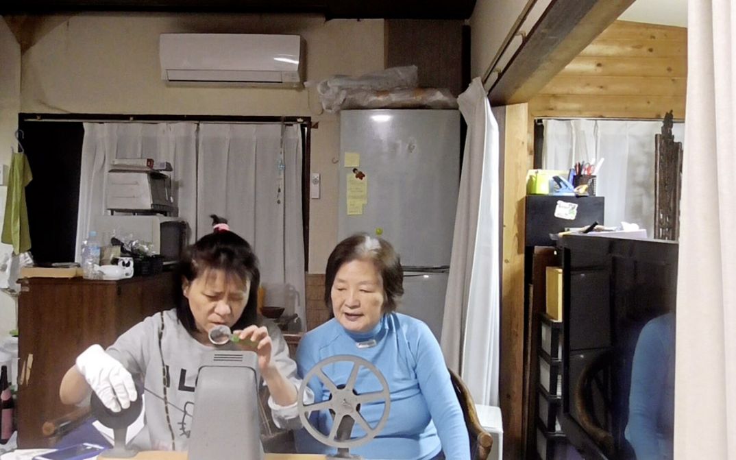 Film still from "Doesarananeun moksori / Yomigaeru Koe" by Park Soo-nam and Park Maeui. It shows two women sitting at a table in a room with a kitchen. The woman on the left is inserting a film into a spool. The woman on the right is watching her.