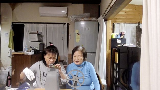 Film still from "Doesarananeun moksori / Yomigaeru Koe" by Park Soo-nam and Park Maeui. It shows two women sitting at a table in a room with a kitchen. The woman on the left is inserting a film into a spool. The woman on the right is watching her. 