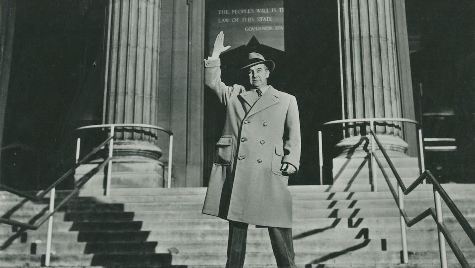 Filmstill aus ALL THE KING'S MEN: Ein Mann steht auf der Treppe eines repräsentativen Gebäudes und hebt seine rechte Hand.
