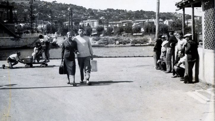 Filmstill aus LA POINTE COURTE: Ein Paar mit einem Koffer geht im Sommer eine Straße in einem Fischerort entlang. Eine Gruppe von Menschen beobachtet sie.