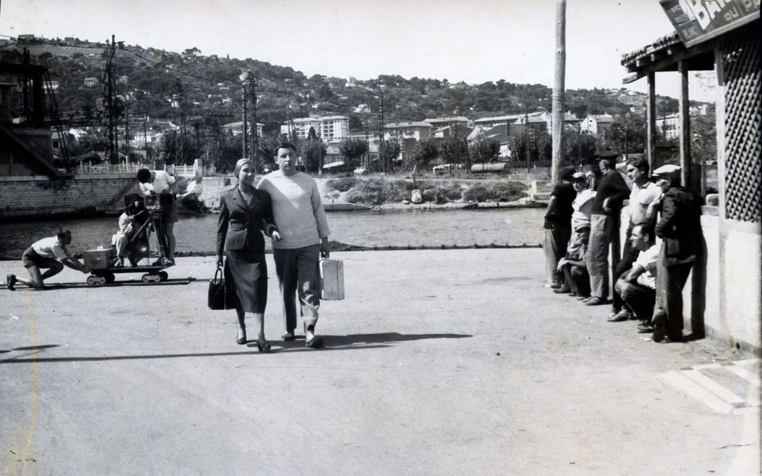 Filmstill aus LA POINTE COURTE: Ein Paar mit einem Koffer geht im Sommer eine Straße in einem Fischerort entlang. Eine Gruppe von Menschen beobachtet sie.